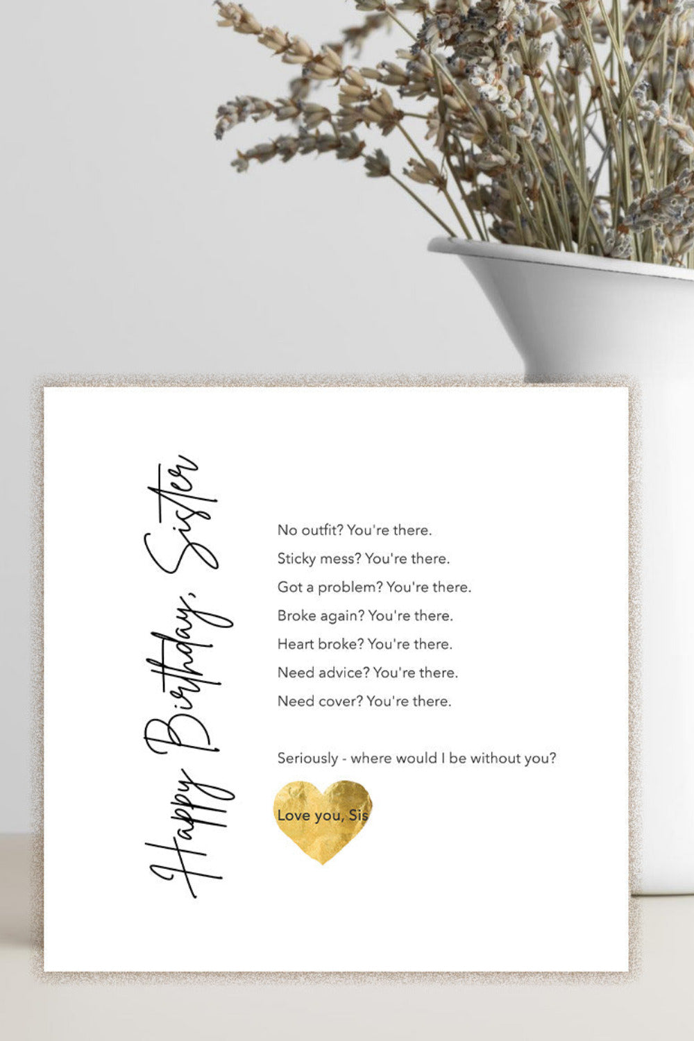 A square white birthday card with black script and poem text, plus a small gold heart design, displayed on a light surface beside a white vase with dried flowers.