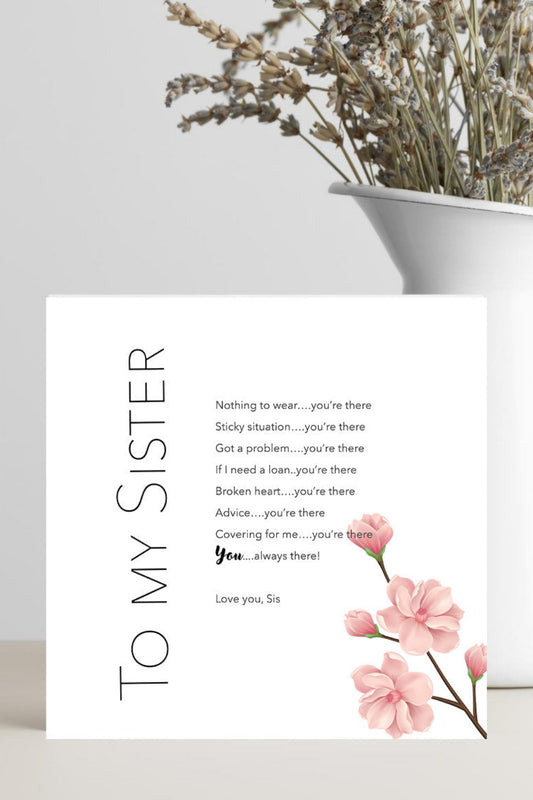 A white greeting card with black text reading 'TO MY SISTER' and a poem, decorated with pink flowers at the bottom, standing on a light surface next to a white vase with dried flowers.