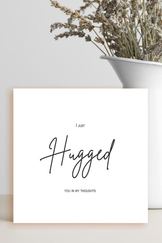 A square white art print with black cursive text reading 'I just Hugged YOU IN MY THOUGHTS' stands on a light beige surface. A white ceramic vase with dried flowers sits behind it against a light gray wall.