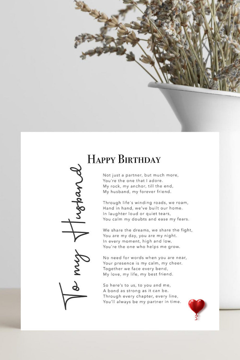 A square white birthday card with a printed poem titled 'Happy Birthday To My Husband' and a small red heart at the bottom, displayed on a light surface beside a white planter with dried flowers.