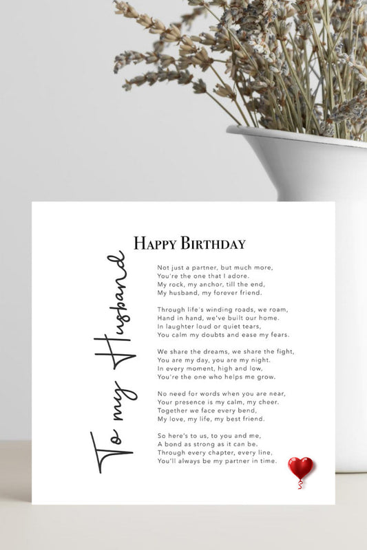 A square white birthday card with a printed poem titled 'Happy Birthday To My Husband' and a small red heart at the bottom, displayed on a light surface beside a white planter with dried flowers.