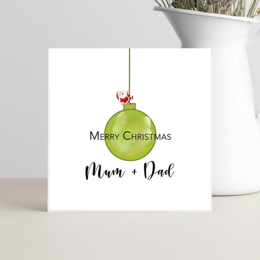 Card with father Christmas climbing a green Christmas ornament and text 'Merry Christmas Mum & Dad' on a white background.  